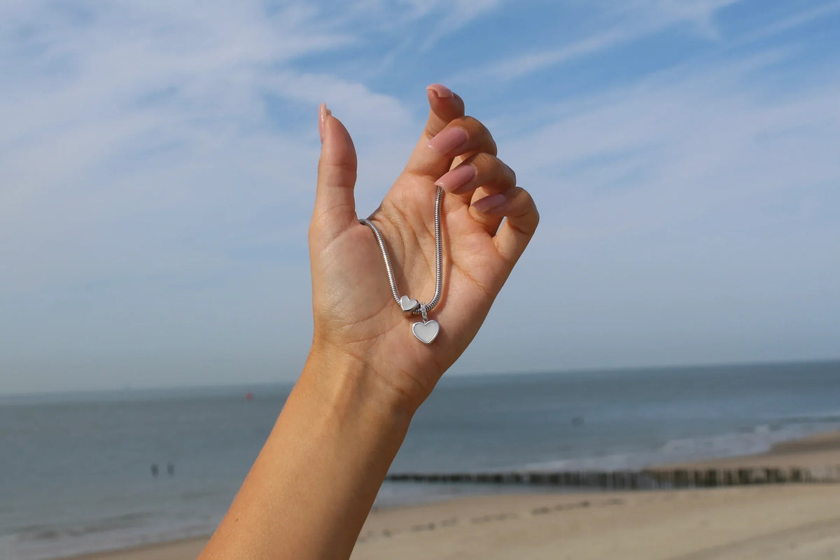 Charm-Armband aus glänzendem Silber mit herzförmigem Anhänger, gehalten vor einer Küstenlandschaft.