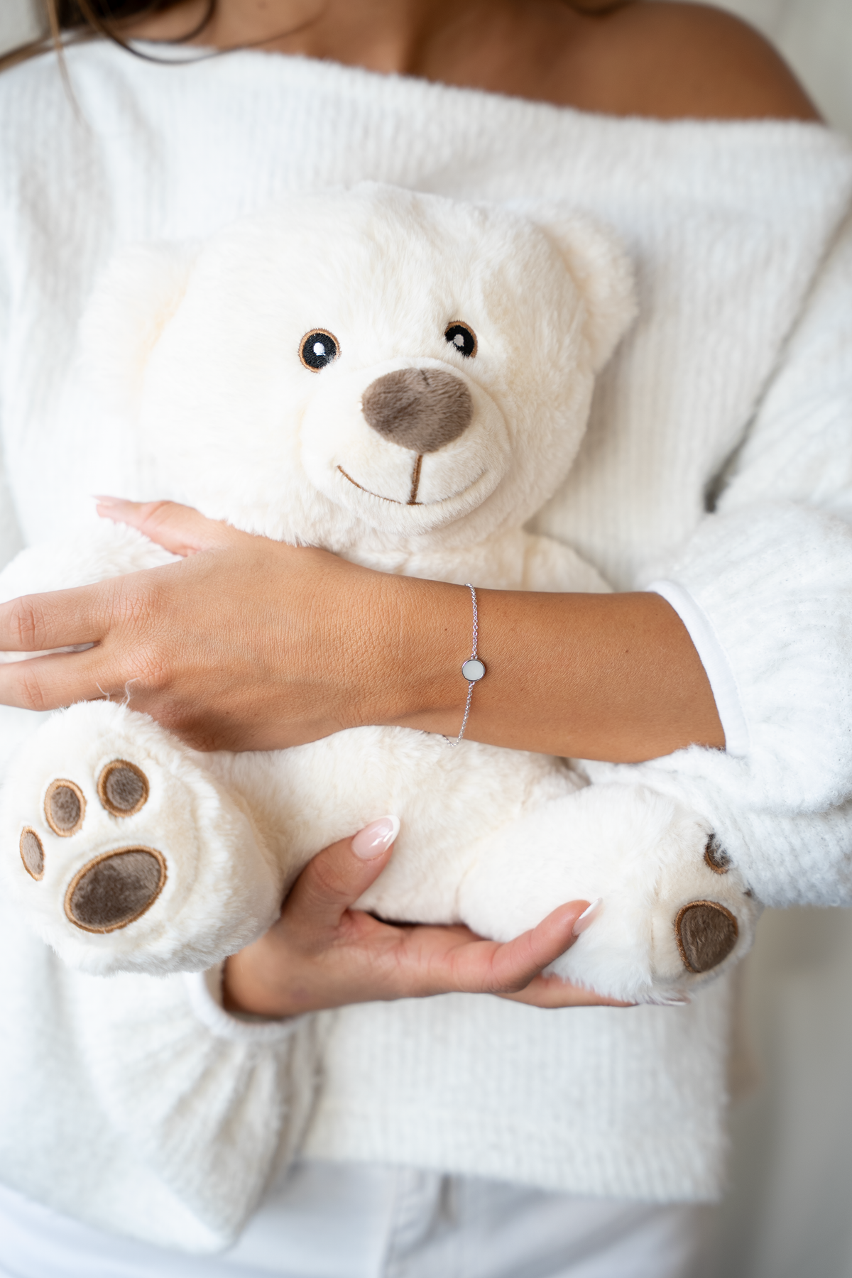 Frau im weißen Pullover umarmt einen Teddybären und trägt das silberne Armband Lis mit rundem Muttermilchstein.