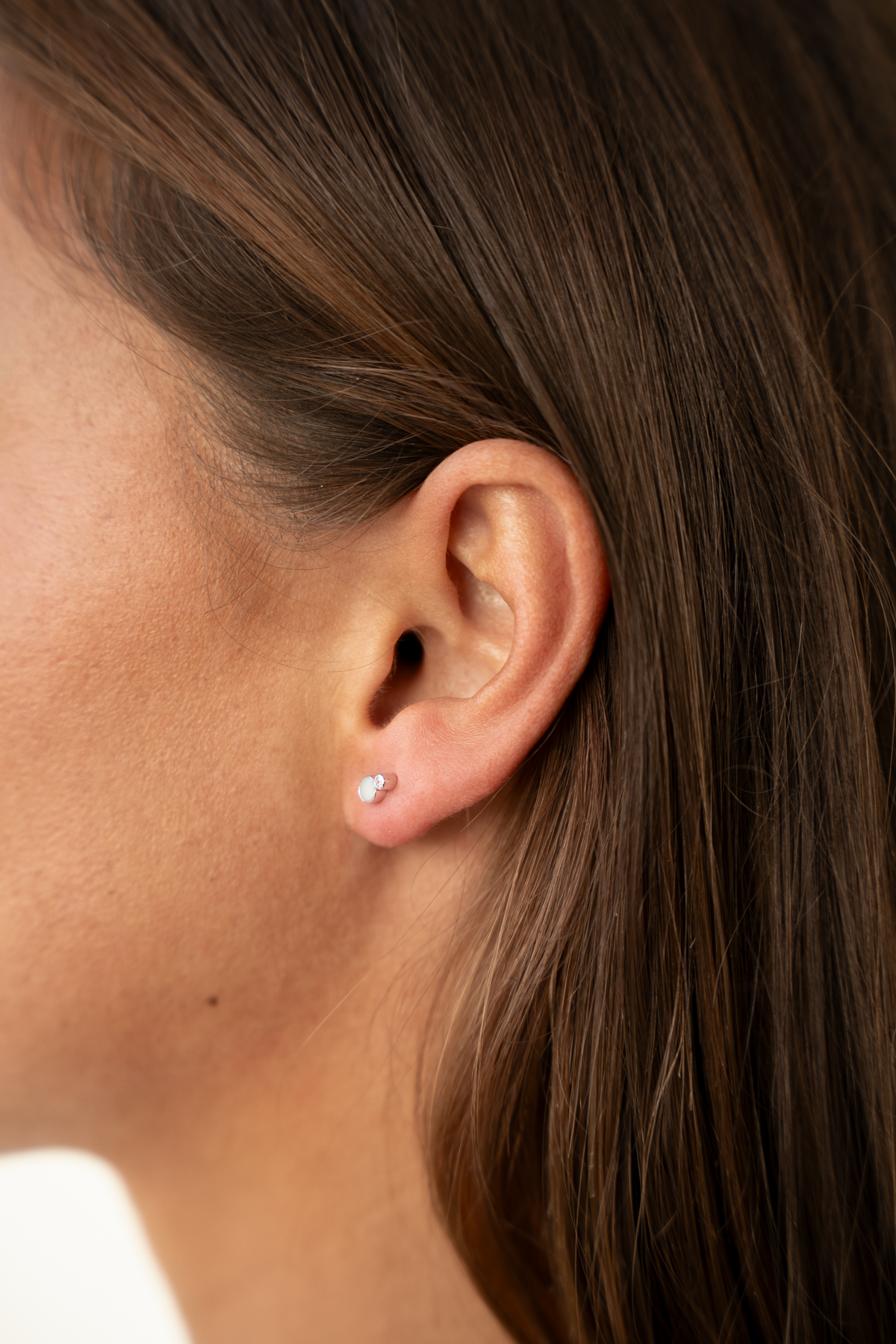 Nahaufnahme eines Ohrläppchens mit kleinem Muttermilch-Ohrstecker, lange braune Haare im Hintergrund